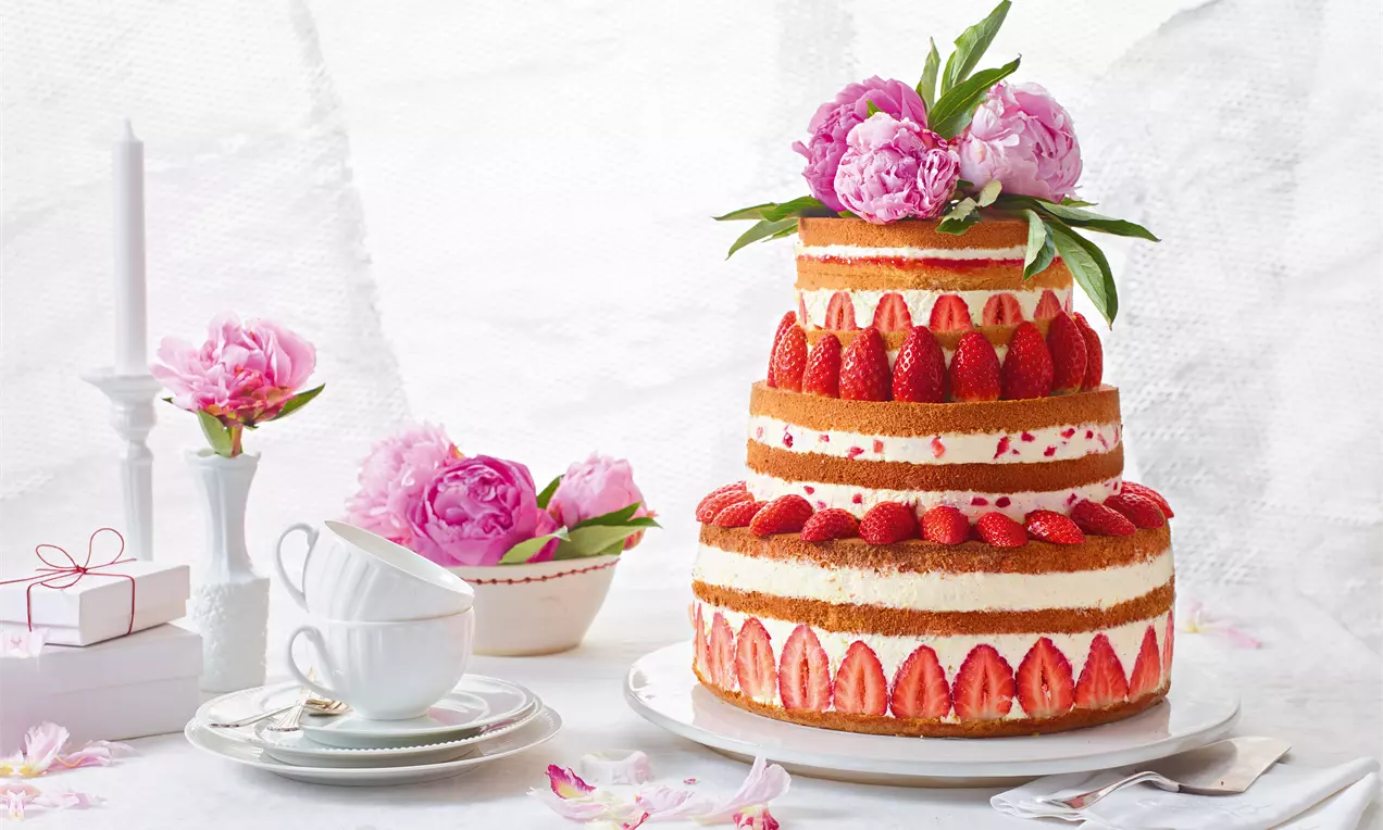 Tort festiv în trei etaje cu blat auriu, straturi de cremă albă, decorat cu căpșuni întregi și felii, flori roz și frunze verzi pe un platou alb