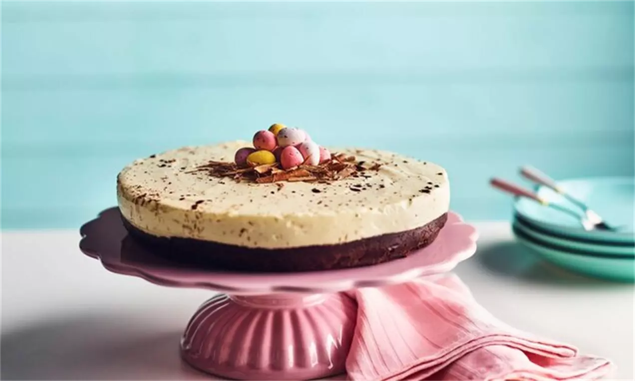 Cheesecake cu ciocolată albă pe blat brun de biscuiți, decorat cu ouă colorate de zahăr și fulgi de ciocolată, așezat pe un platou roz
