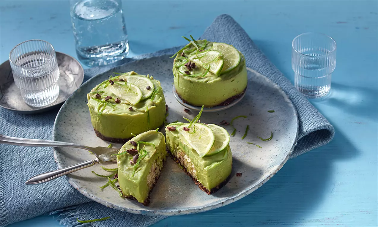 Mini prăjituri raw cu cremă verde de avocado și lime pe blat crocant, decorate cu felii de lime, coajă rasă și fulgi de cacao pe o farfurie în nuanțe de bleu