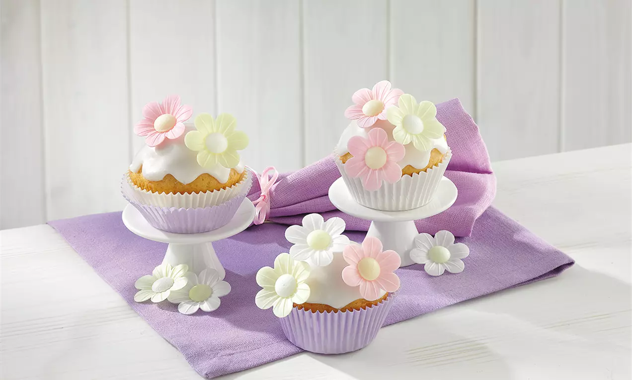 Brioșe vegane aurii, glazurate alb, înformate în hârtii mov pastel, decorate cu flori delicate din zahăr roz și alb, așezate pe suporturi albe
