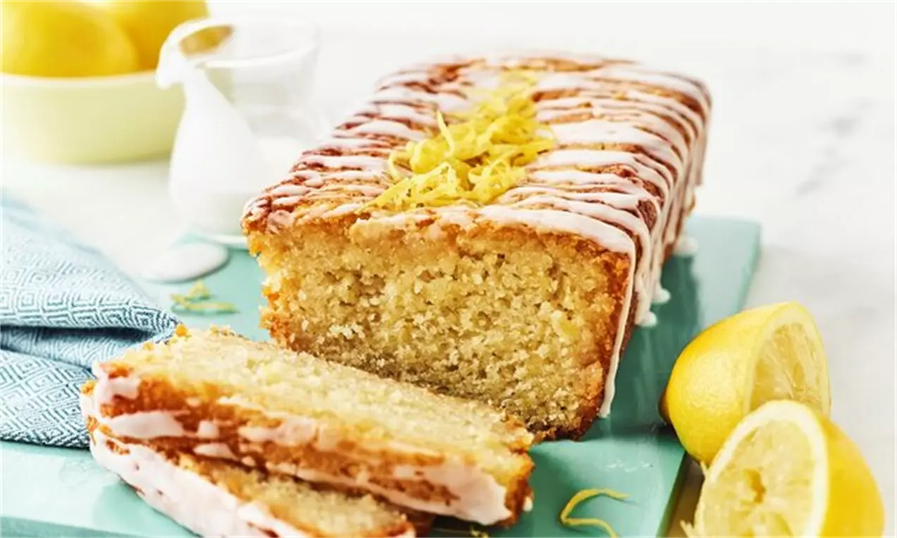 Chec vegan cu lămâie, galben-deschis, glazurat cu dungi albe și decorat cu coajă de lămâie pe un platou turcoaz, alături de felii de lămâie