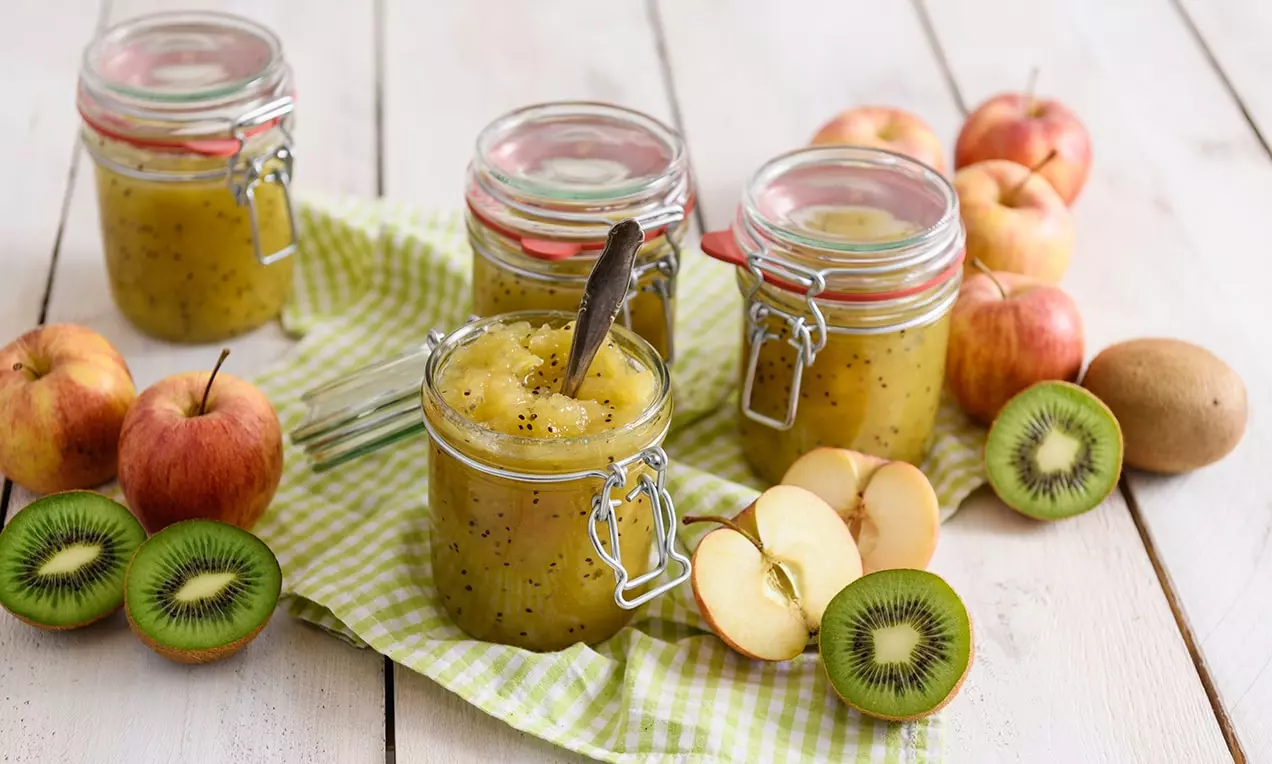 Dulceață aurie de mere și kiwi cu semințe mici negre, așezată în borcane de sticlă pe un șervet verde în dungi, lângă felii de fructe proaspete