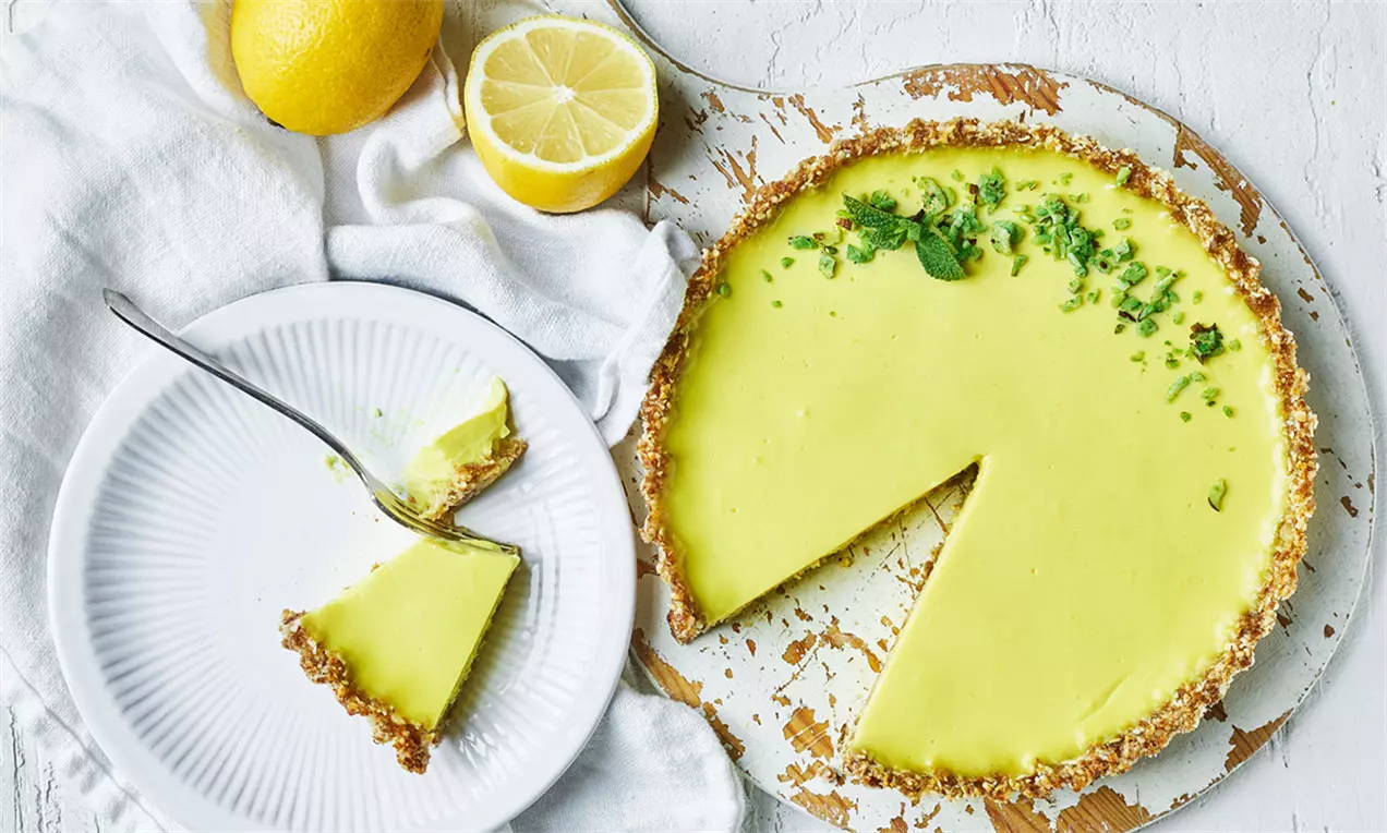 Tartă vegană cu lămâie în nuanțe vibrant galben-verzui, cu blat crocant și decor de frunze proaspete, pe un platou rustic alb