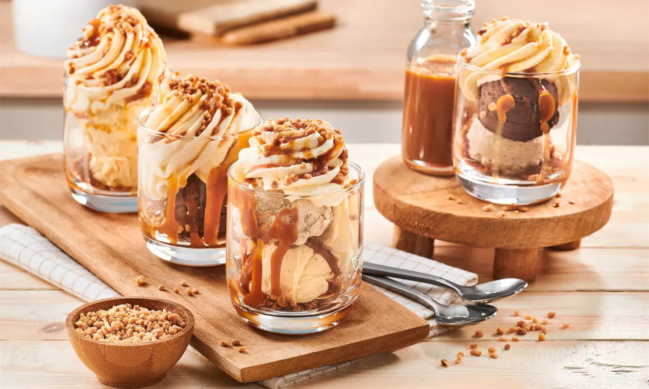 Înghețată de banane și cacao în pahare transparente, acoperită cu caramel auriu, frișcă spiralată și granule crocante pe o masă din lemn