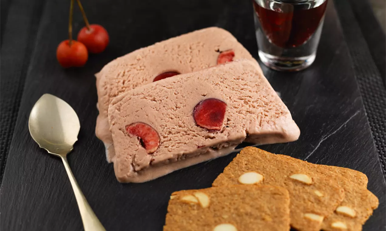 Feliile de semifreddo roz-pal cu cireșe întregi înglobate, alături de biscuiți aurii cu migdale pe un platou de ardezie neagră