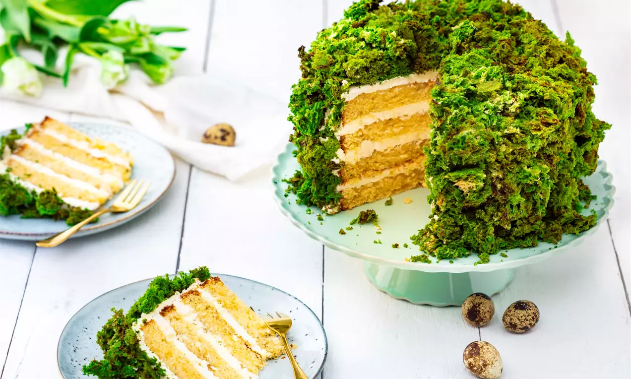 Tort verde vibrant cu blat stratificat galben-auriu și cremă albă, decorat cu mușchi de copac pe un suport pastel pe masă albă