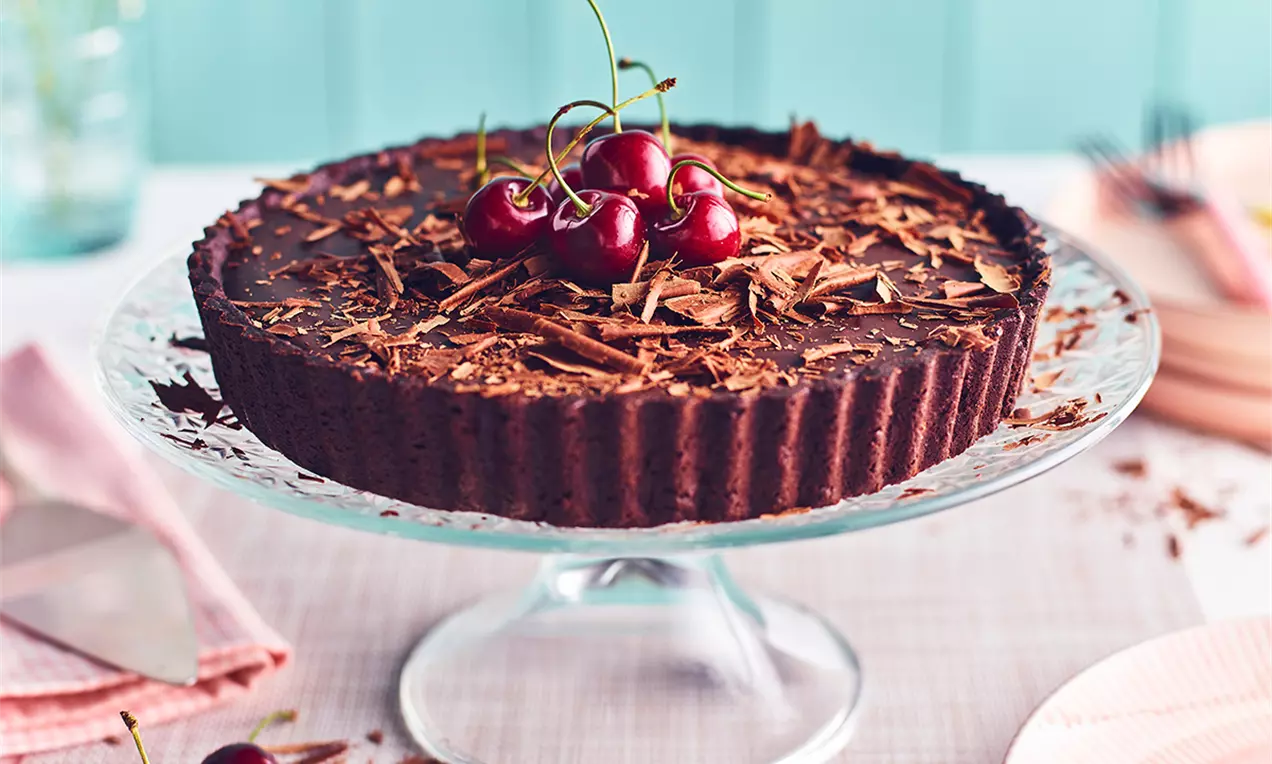 Tartă cu crustă dantelată din cacao, cremă lucioasă de ciocolată extra neagră, decorată cu cireșe proaspete și fulgi de ciocolată pe un platou transparent