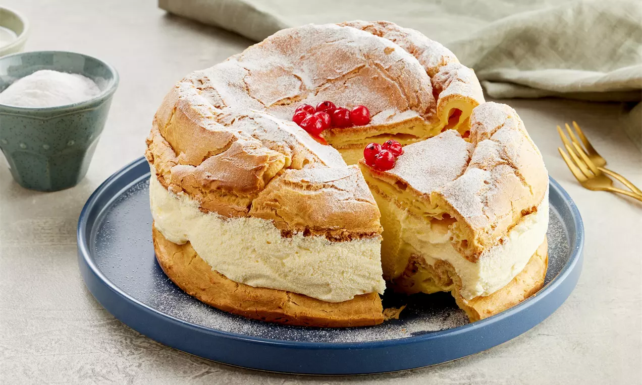 Tort ecler cu straturi aurii pufoase, cremă albă delicată, decorat cu zahăr pudră și bobițe roșii pe un platou albastru