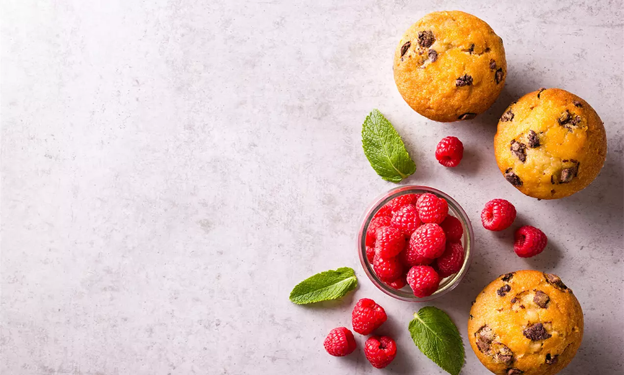 Cupcakes cu vanilie aurii și bucăți de ciocolată, alături de zmeură roșie proaspătă și frunze verzi pe o farfurie albă subtilă