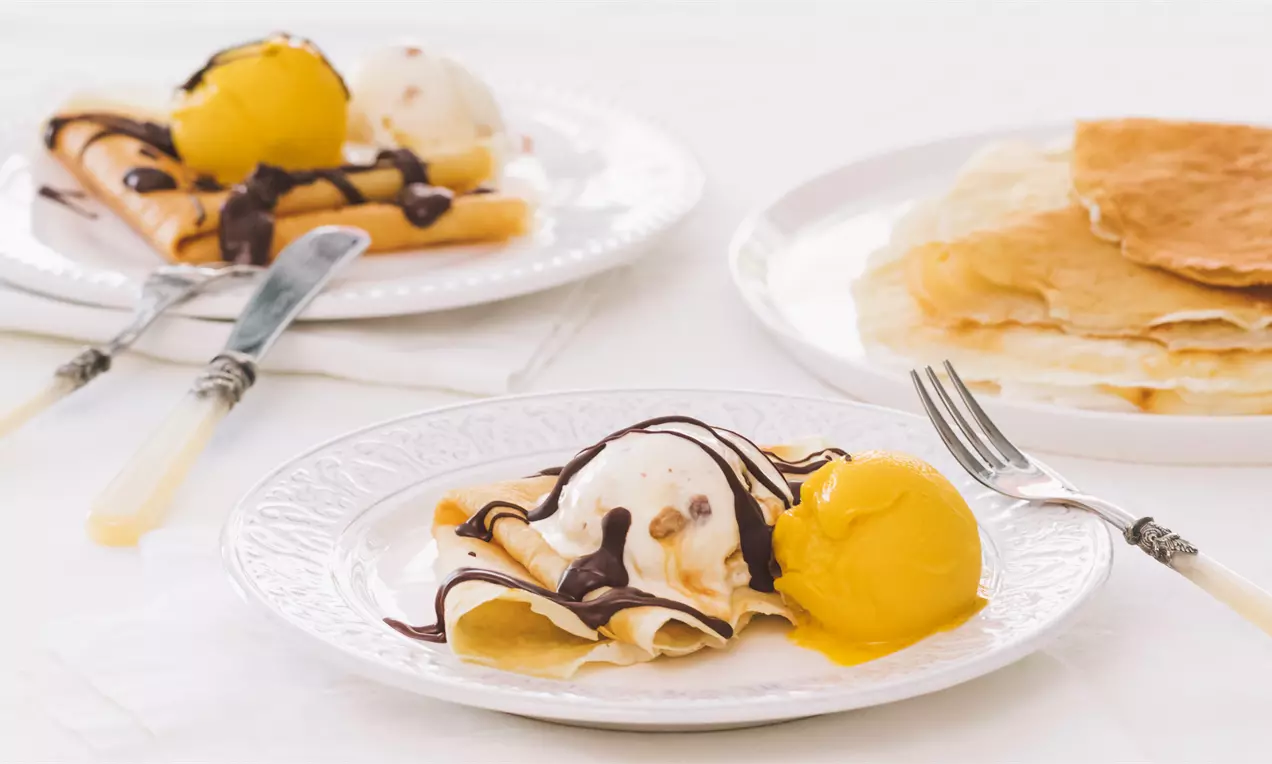 Înghețată de mango galben-strălucitor alături de un glob de înghețată vanilie pe clătite aurii stropite cu sos de ciocolată pe o farfurie albă