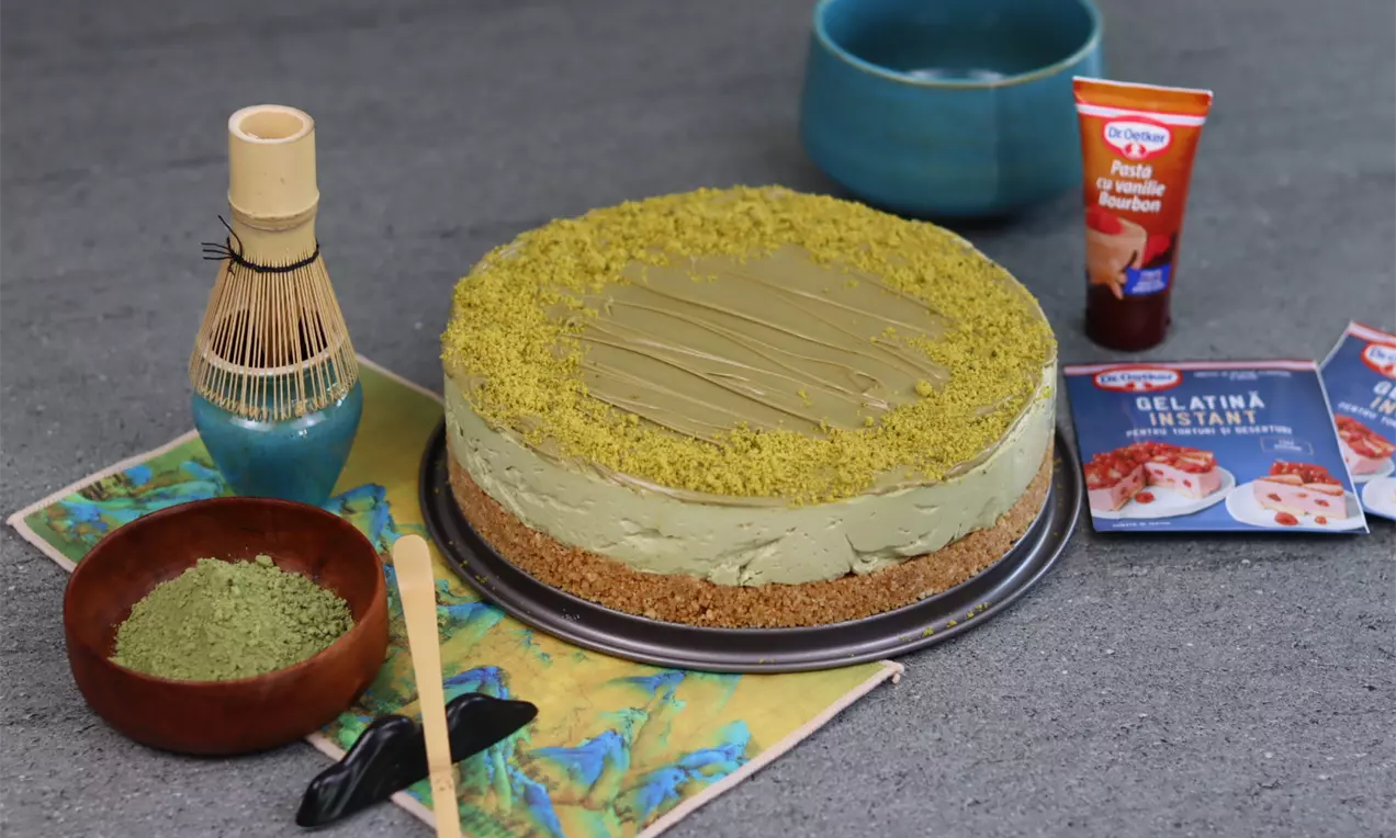 Cheesecake cu blat crocant maro, umplutură cremoasă verde-pastel de matcha, decorat cu pudră fină de fistic și cremă uniform întinsă