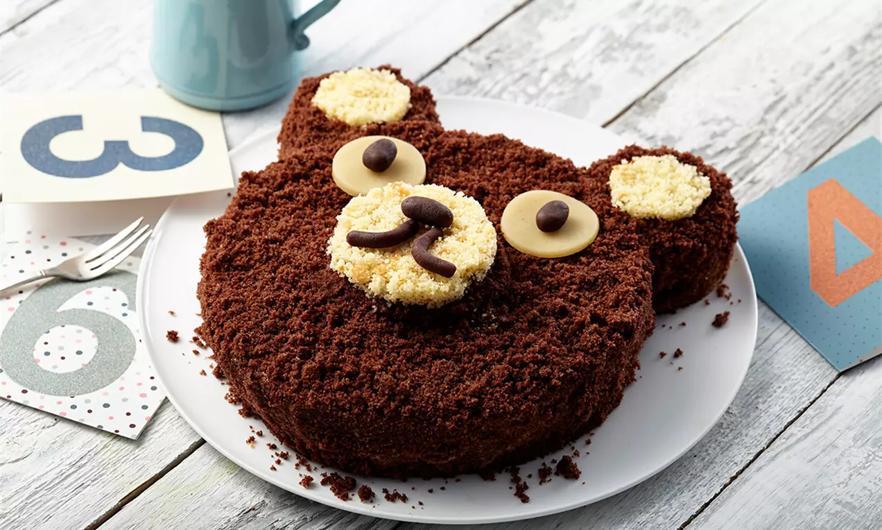 Tort în formă de ursuleț cu blat sfărâmicios de cacao, urechi și bot decorate cu cremă deschisă la culoare și detalii din ciocolată