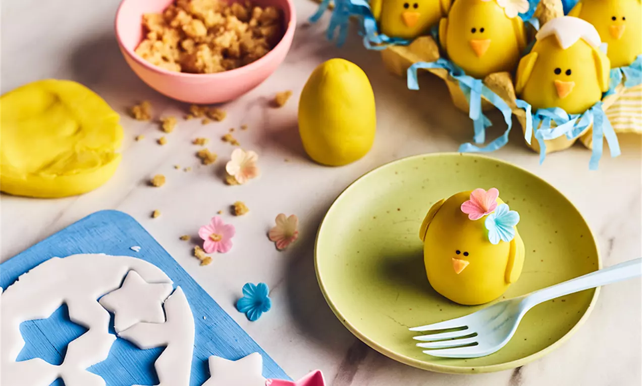 Cake pops în formă de puișori galbeni de Paște, cu ochi negri, cioc portocaliu și decorațiuni florale, așezați pe farfurii și paie albastre