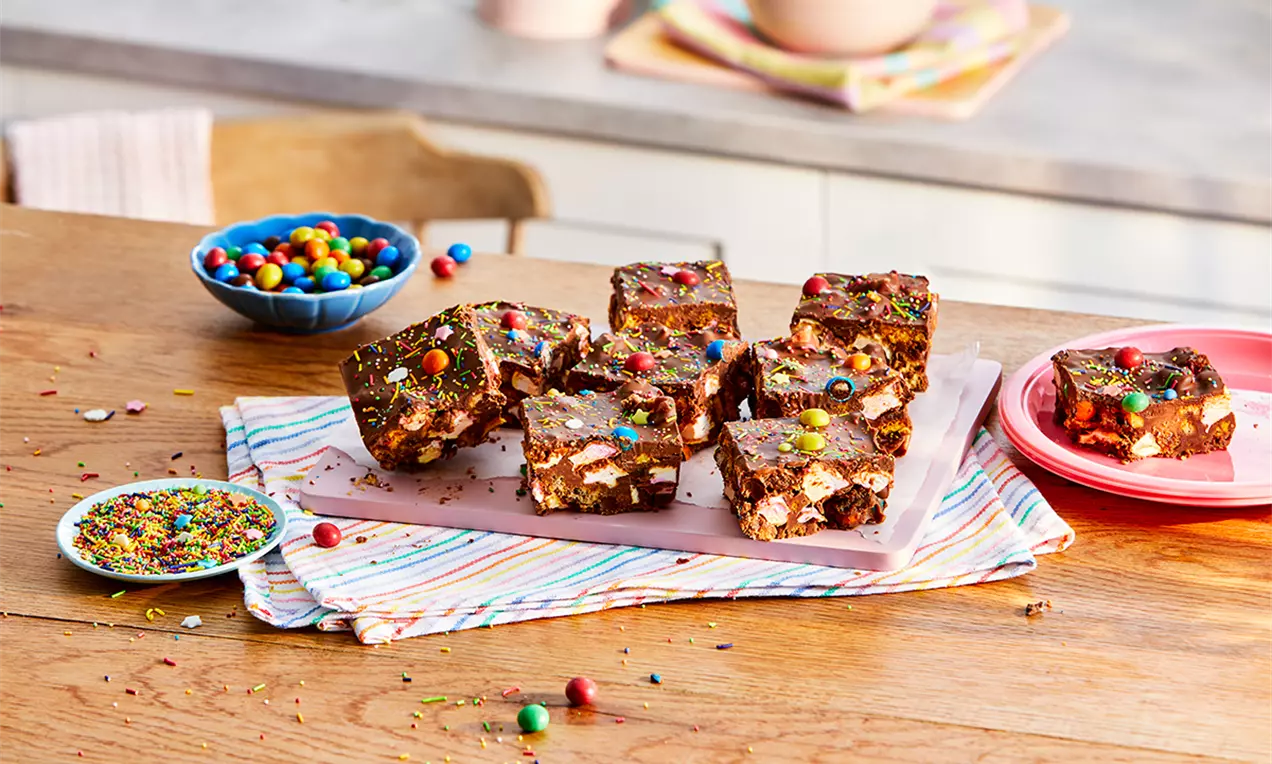 Cuburi de Rocky Road cu textură densă de ciocolată, bucăți de bezea și nuci, presărate cu bombonele colorate pe un platou roz