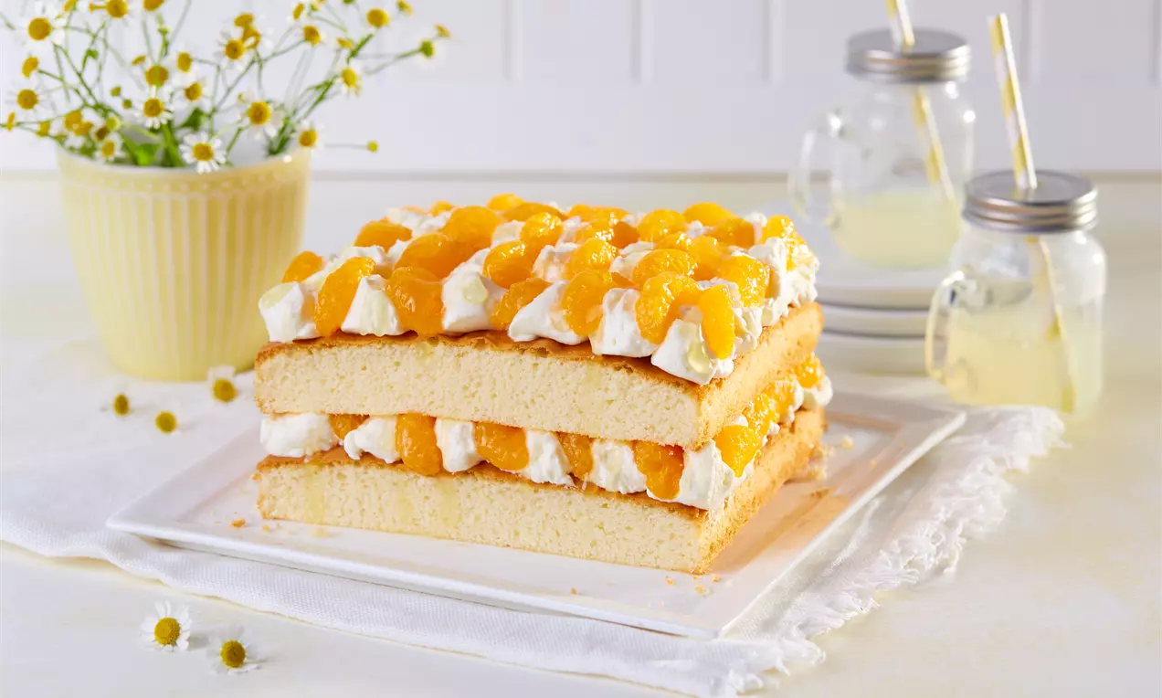 Tort stratificat cu blat pufos bej, frișcă albă cremoasă și bucăți de mandarine portocalii, așezat pe o farfurie albă într-un decor luminos