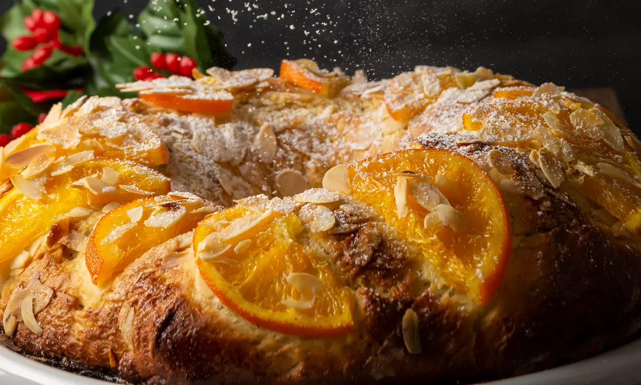 Coroniță aurie din aluat pufos decorată cu felii de portocală caramelizate, fulgi de migdale și zahăr pudră, pe un fundal festiv
