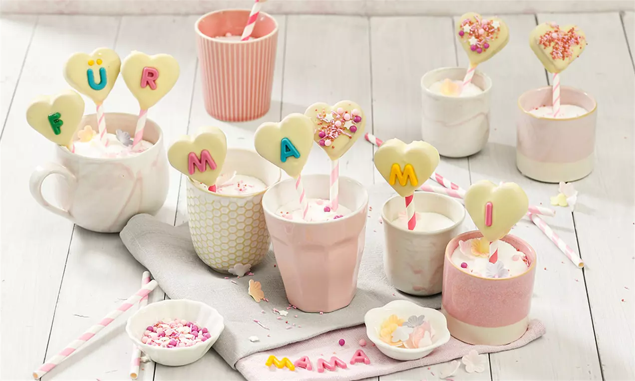Prăjituri-acadele în formă de inimă din ciocolată albă, decorate cu litere colorate și bombonele, în pahare delicate cu nuanțe pastelate