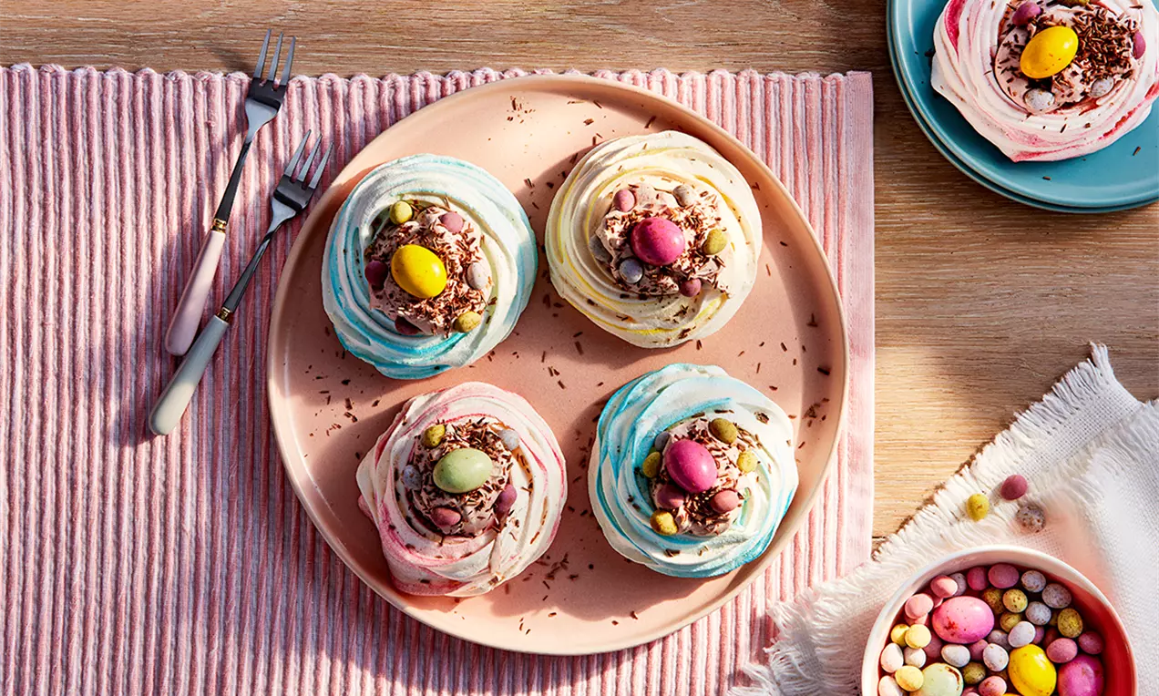 Cuiburi de bezea pastelate, albe și roz cu margini albastre, gălbenușuri colorate și fulgi de ciocolată pe o farfurie roz pe masă decorată cu bomboane