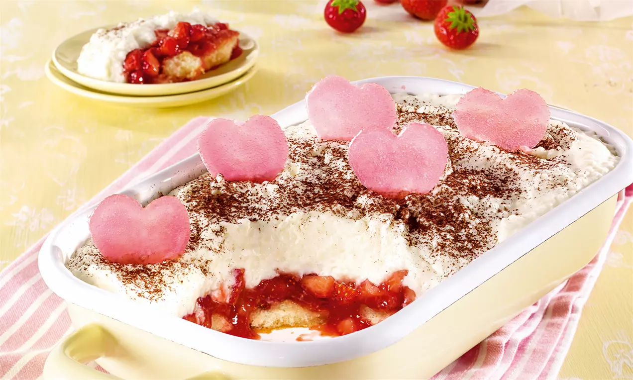 Desert cremos stratificat cu textură aerată albă, bucăți roșii de căpșuni și decor din inimioare roz pe un vas dreptunghiular alb