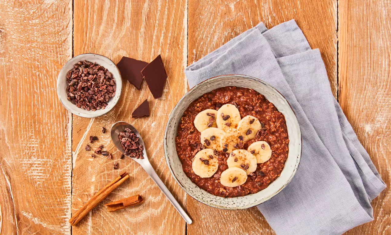 Bol ceramic gri umplut cu fulgi de ovăz cu ciocolată, decorat cu felii de banană, fulgi de cacao și scorțișoară, pe masă din lemn rustic