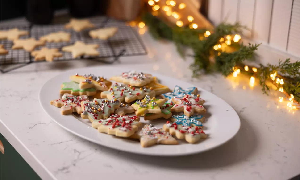 Biscuiți de Crăciun în formă de stea, glazurați în culori vibrante și presărați cu decoruri colorate, așezați pe un platou alb pe blat marmorat