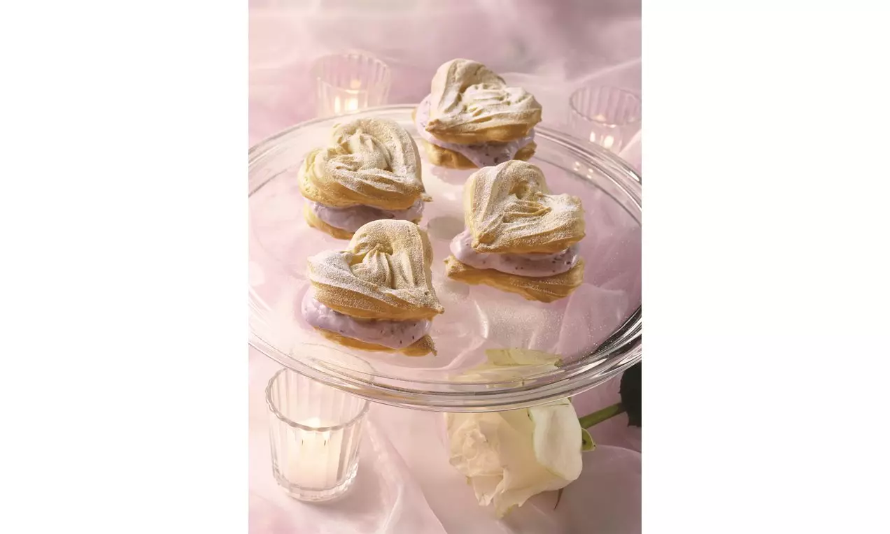 Biscuiți inimioare aurii, cu umplutură cremă lila, acoperiți cu glazură fină, așezați pe un platou transparent cu decor romantic de lumânări
