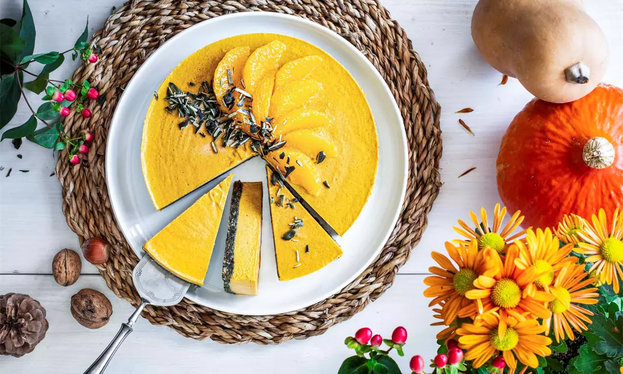 Plăcintă vegană cu dovleac, strat neted portocaliu intens, decorată cu felii de dovleac și semințe pe o farfurie albă, așezată pe suport împletit
