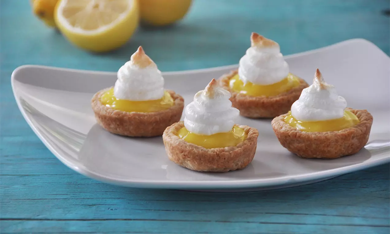 Mini tarte aurii cu crustă crocantă, umplute cu cremă de lămâie galben intens și bezea pufoasă rumenită, aranjate pe o farfurie albă
