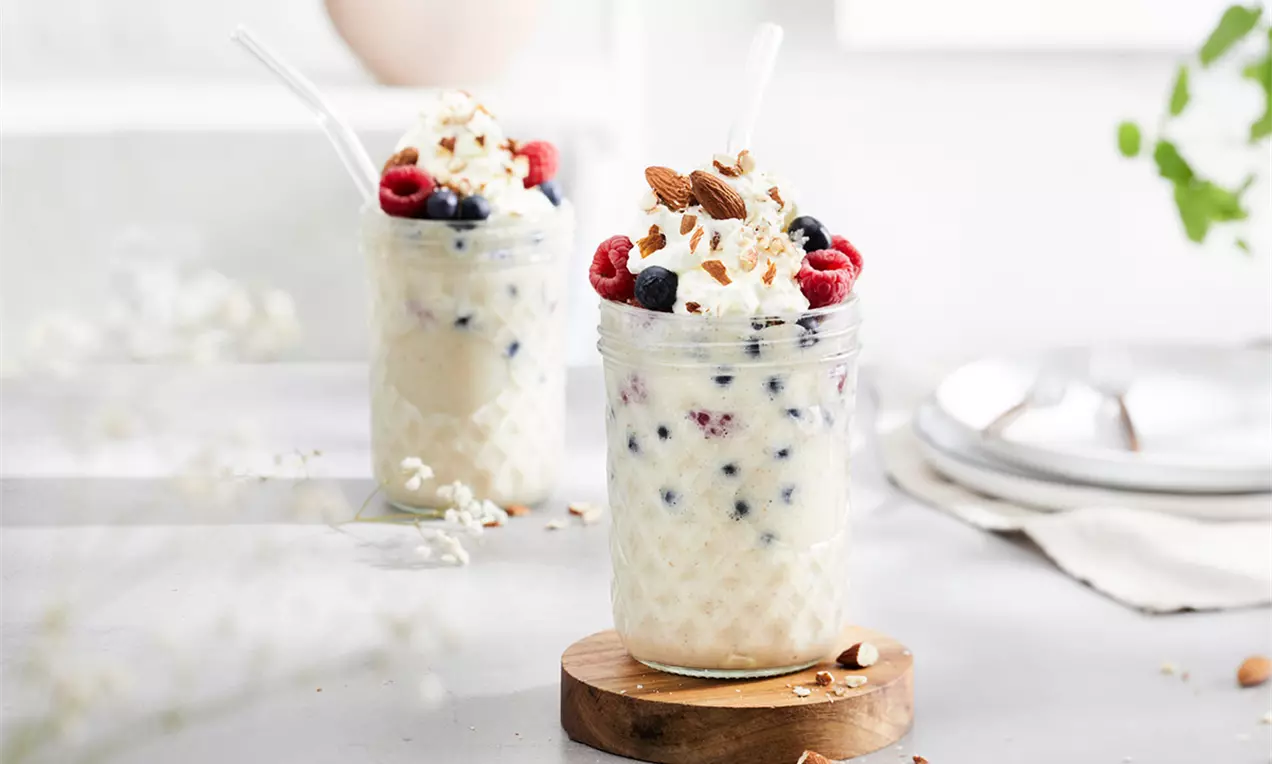 Milkshake vegan cremos, alb-vanilat, cu fructe de pădure, frișcă, migdale crocante și pai alb, servit într-un borcan pe un suport de lemn