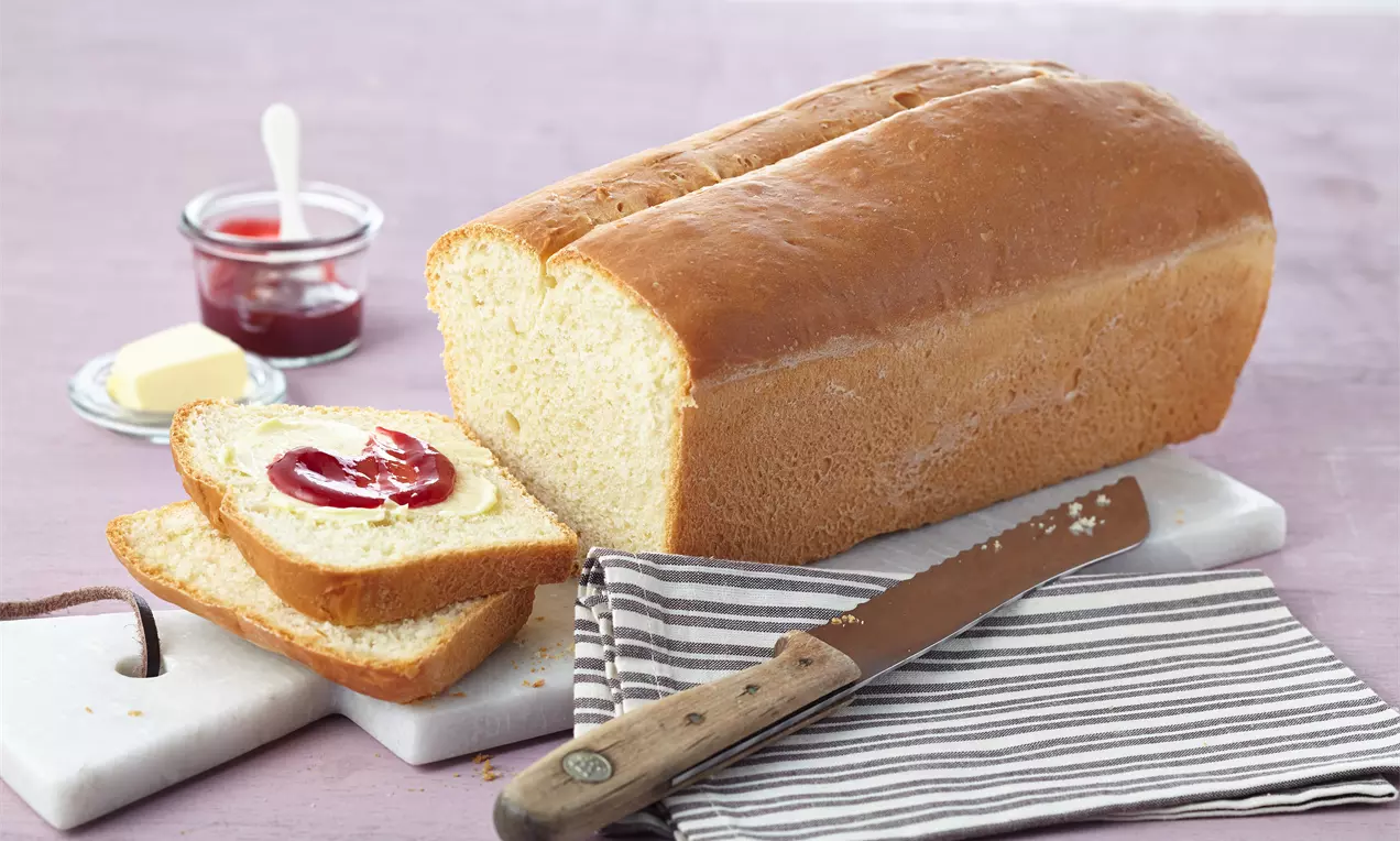 Pâine albă pufoasă, cu coajă aurie uniform rumenită, feliată alături de unt, gem roșu și un cuțit de lemn pe o masă pastelată