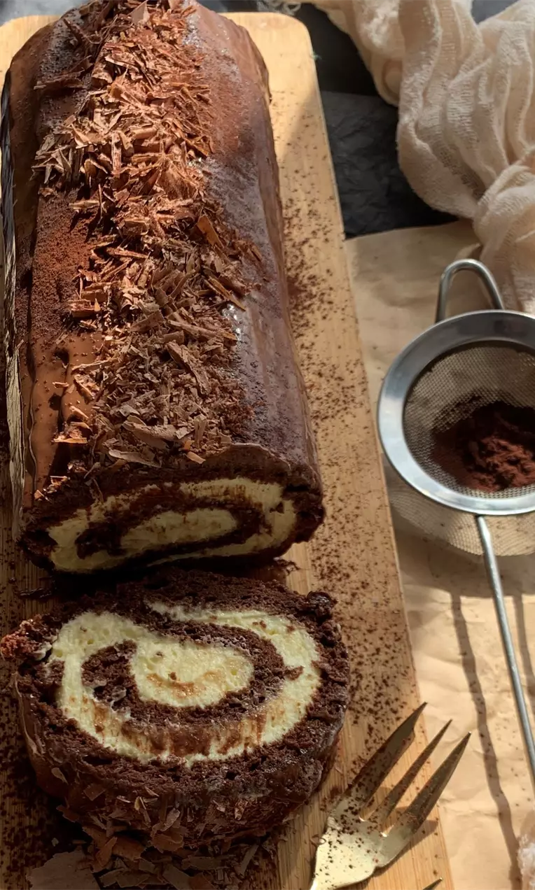 Ruladă cu ciocolată lucioasă decorată cu fulgi de ciocolată pe un platou de lemn, cu secțiune ce dezvăluie spirale vanilie și ciocolată