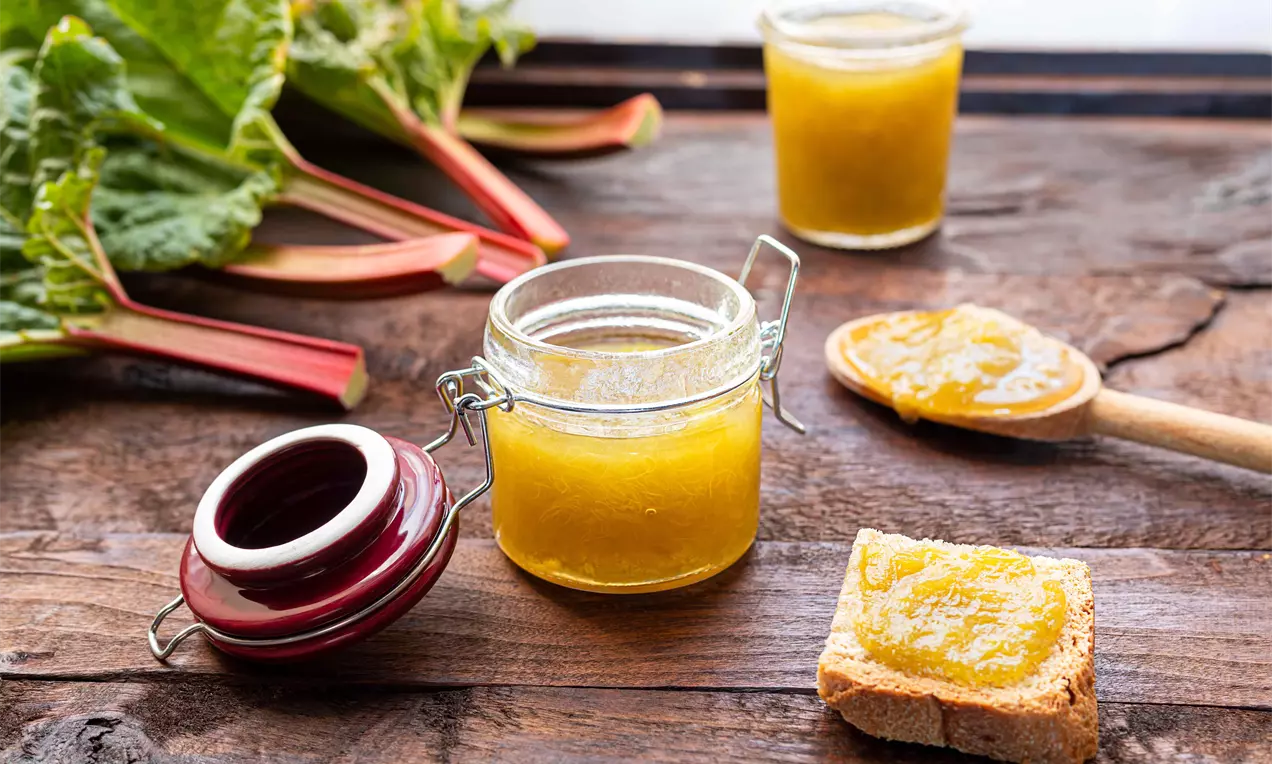 Dulceață aurie de rubarbă și mango în borcan de sticlă, întinsă pe o felie de pâine și pe o lingură de lemn, alături de tulpini de rubarbă