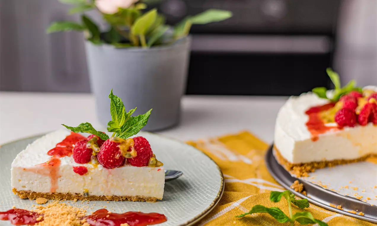 Cheesecake de vanilie fără coacere cu textură fină și strat crocant de biscuiți, decorat cu zmeură, sos roșu și frunze proaspete de mentă pe o farfurie albă