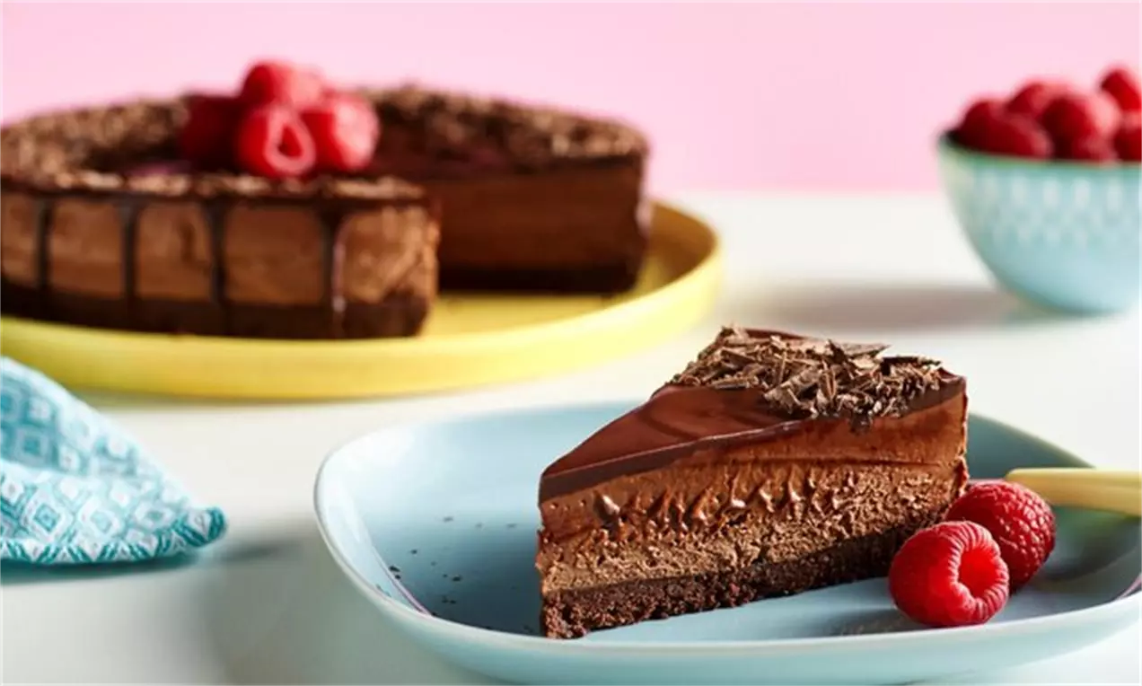 Felie de cheesecake cu ciocolată lucioasă și straturi catifelate maro, ornată cu fulgi de ciocolată și zmeură proaspătă pe o farfurie bleu