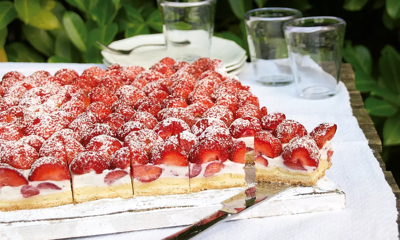 Tort pătrat cu ricotta cremoasă, acoperit cu căpșuni proaspete și zahăr pudră pe un platou alb, pe o masă decorată simplu în aer liber
