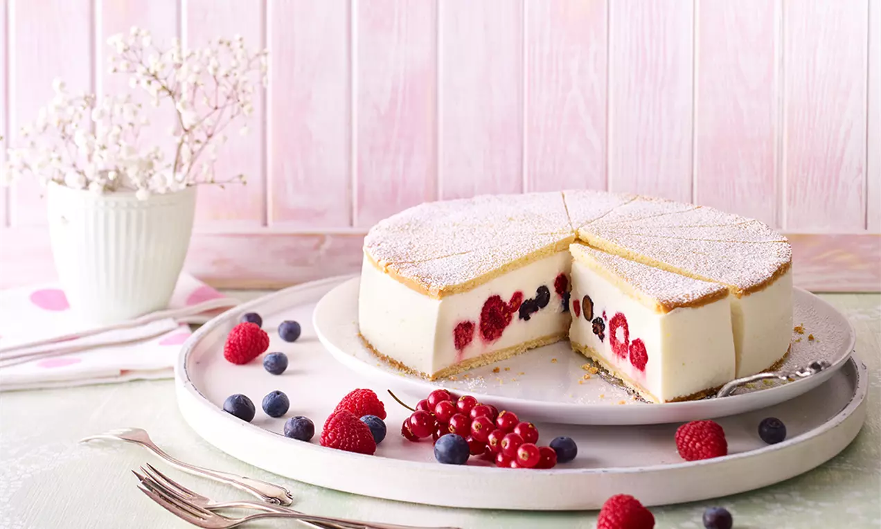 Prăjitură rotundă cu brânză dulce, umplutură albă fină, fructe de pădure colorate și blat subțire, presărată cu zahăr pudră, aranjată pe o farfurie albă