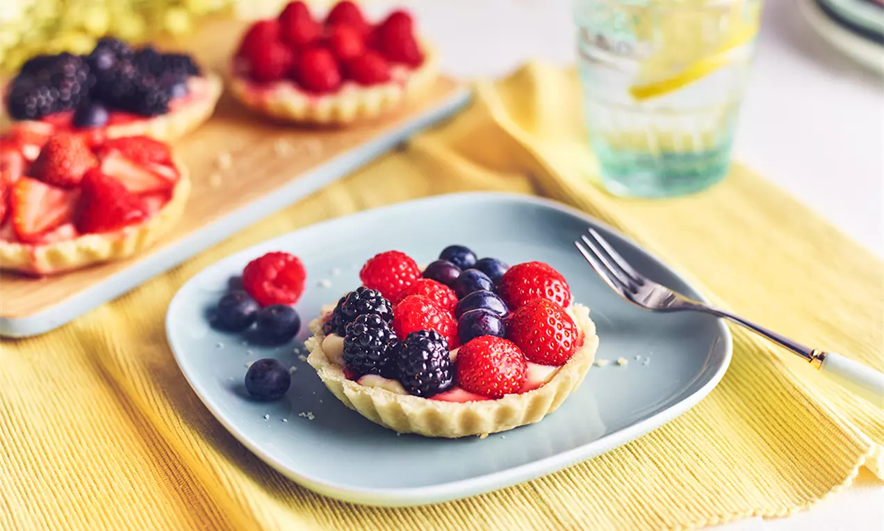Mini tarte cu cremă de vanilie, decorate cu fructe de pădure vibrante, așezate pe o farfurie bleu, pe un șervet galben luminos