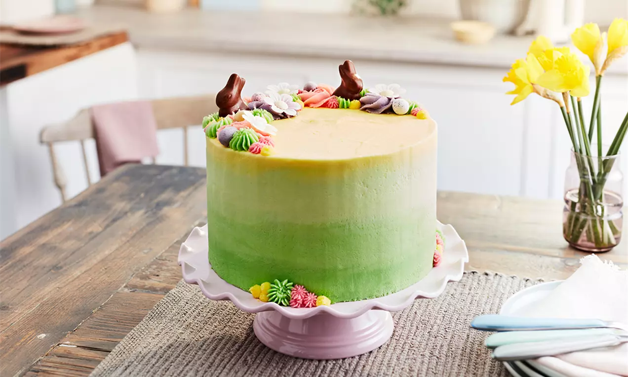 Tort de Paște pe un platou roz, decorat cu glazură degrade galben-verde, flori colorate din cremă și figurine de ciocolată