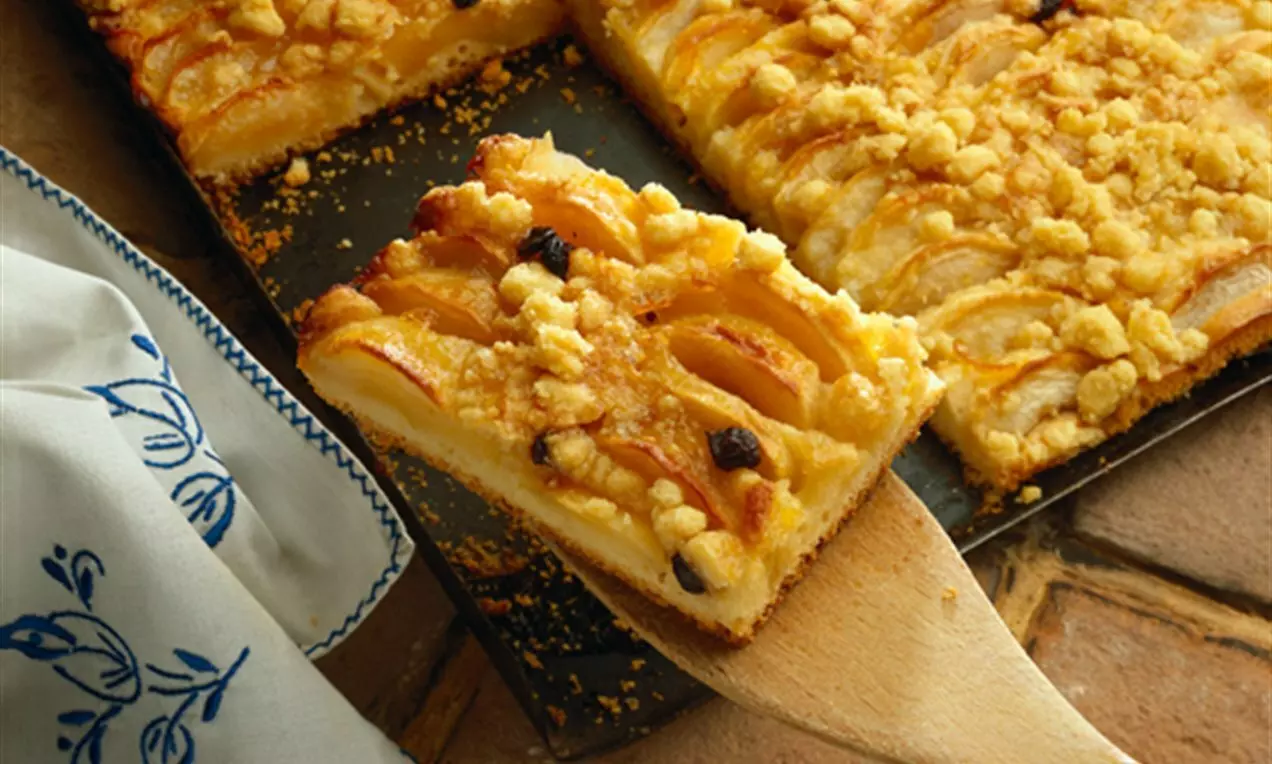 Plăcintă cu mere aurii tăiată în pătrate, cu fâșii de mere așezate simetric, presărată cu crumble crocant pe un fundal rustic