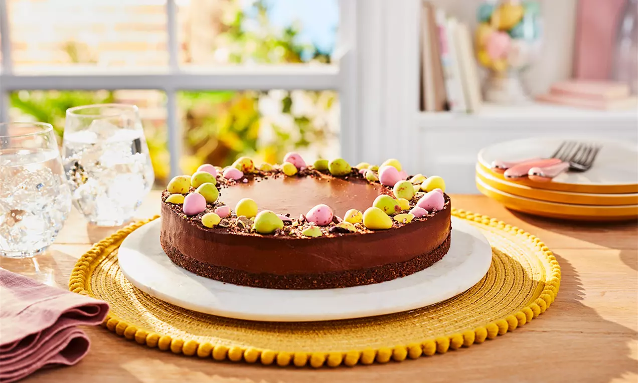 Cheesecake vegan de Paște cu glazură lucioasă de ciocolată, decorat cu ouă colorate și crumble crocant pe un platou alb, așezat pe un suport galben