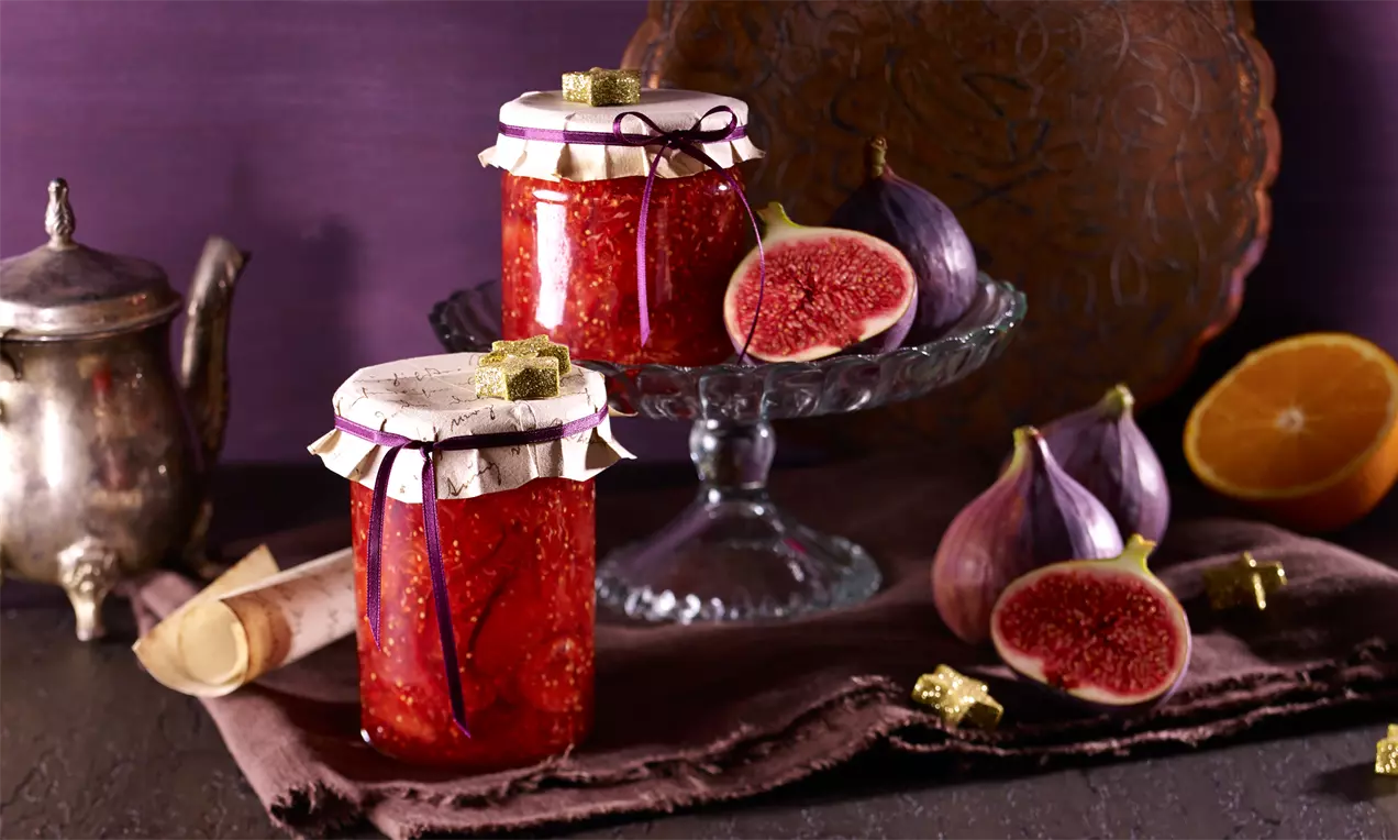 Borcan de dulceață roșie cu capac decorativ lângă smochine mov cu miez roșu și portocală secționată, pe o masă cu fundal violet