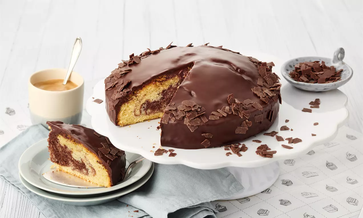 Tort marmorat cu glazură lucioasă de ciocolată maro închis, decorat cu fulgi subțiri de ciocolată, pe un platou alb, alături de o felie servită pe farfurie și o ceșcuță de cafea crem pe un șervet bleu
