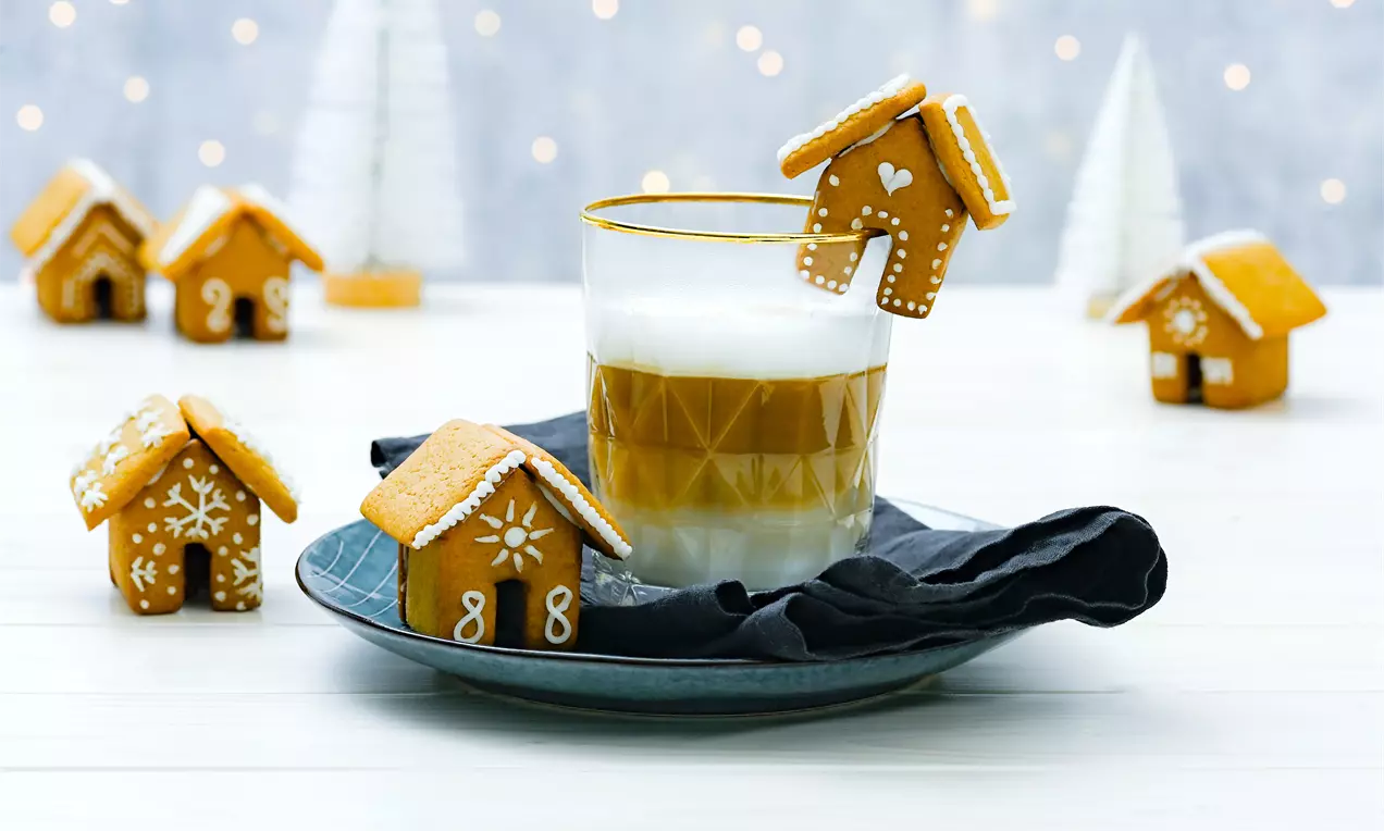 Căsuțe mici de turtă dulce aurii, decorate cu glazură albă, așezate pe un platou luminos alături de un pahar cu strat de latte cremos