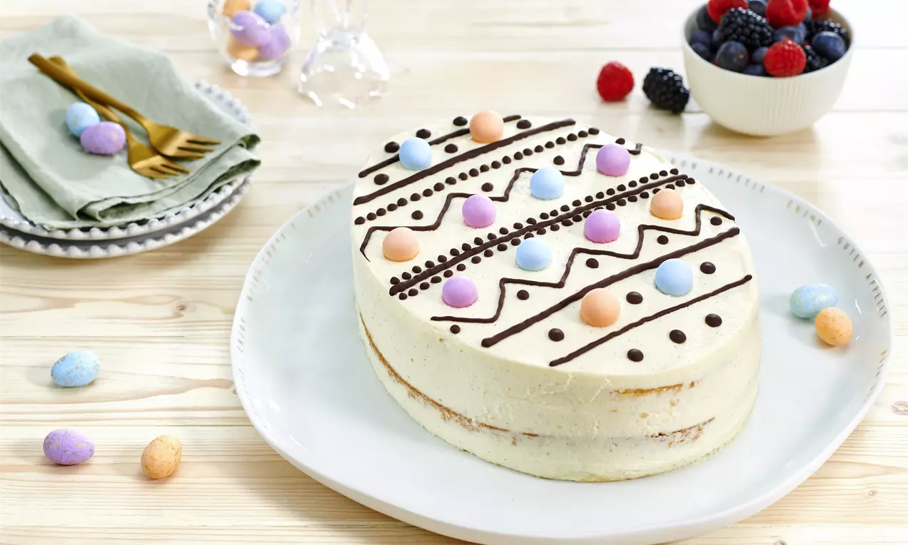 Tort în formă de ou de Paște acoperit cu cremă albă netedă, decorat cu dungi de ciocolată și bombonele colorate pe o farfurie albă