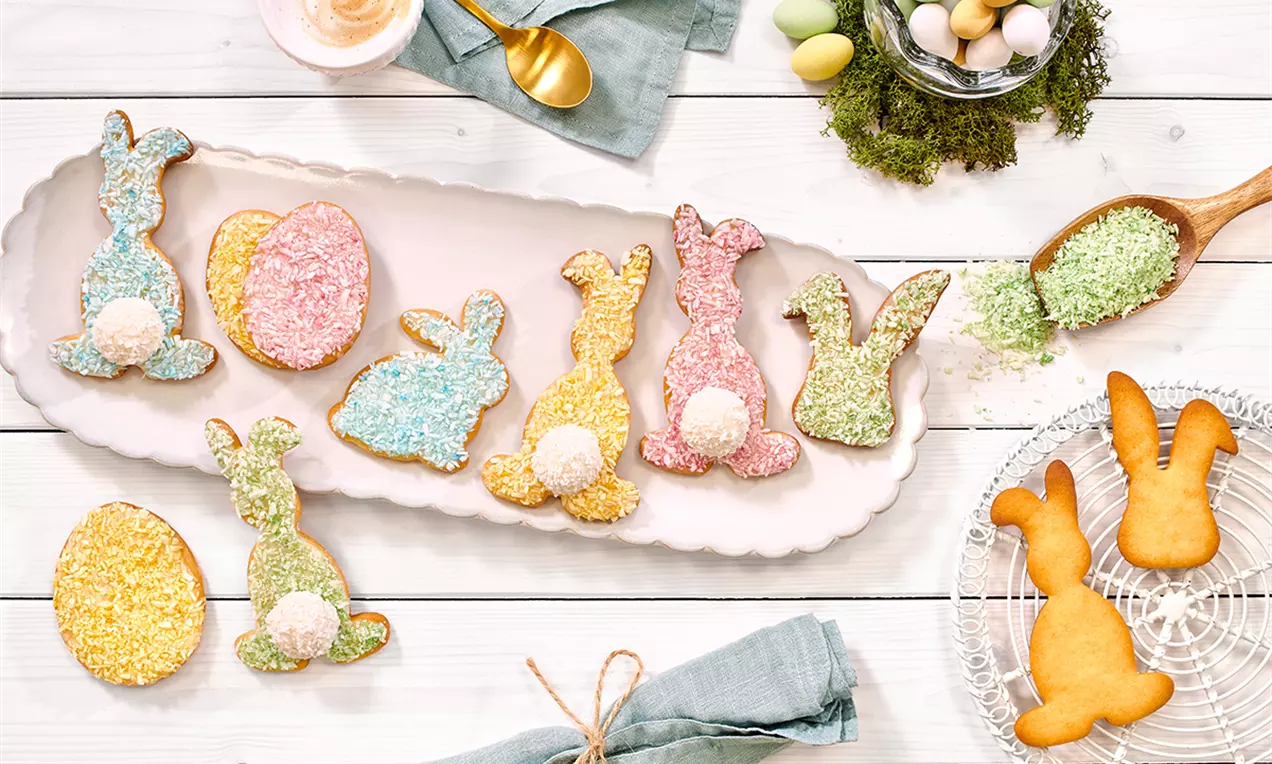 Biscuiți în formă de iepurași și ouă, acoperiți cu nucă de cocos colorată roz, galben, albastru și verde, aranjați pe o tavă albă
