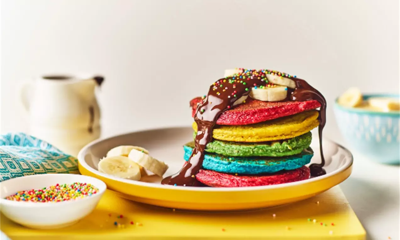 Clătite în straturi viu colorate de roșu, galben, verde și albastru, cu topping de ciocolată și felii de banană pe o farfurie albă