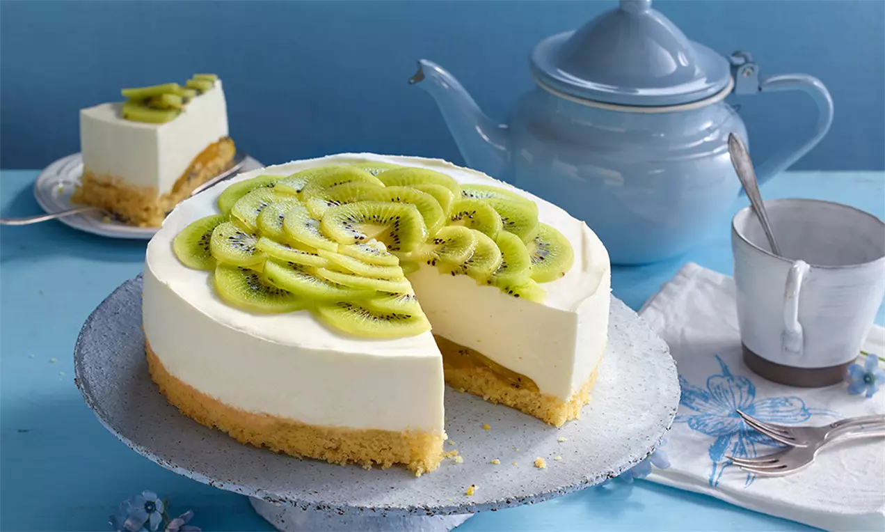 Prăjitură circulară cu cremă albă fină, blat auriu și strat de felii subțiri de kiwi verde deschis pe un platou alb pe fundal albastru