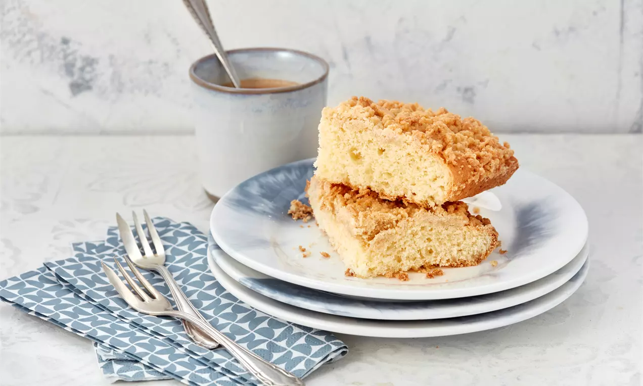 Prăjitură Streusel aurie, cu strat crocant de crumble și textura pufoasă, așezată pe farfurii albe lângă un șervețel albastru și un pahar cu cafea