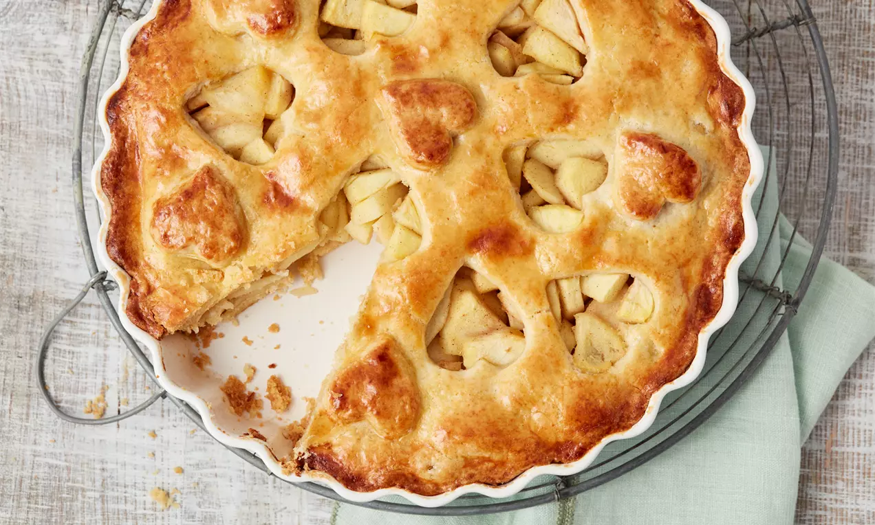 Plăcintă aurie cu mere, cu aluat fraged decorat cu decupaje în formă de inimă, aranjată într-o tavă albă pe un suport metalic rotund
