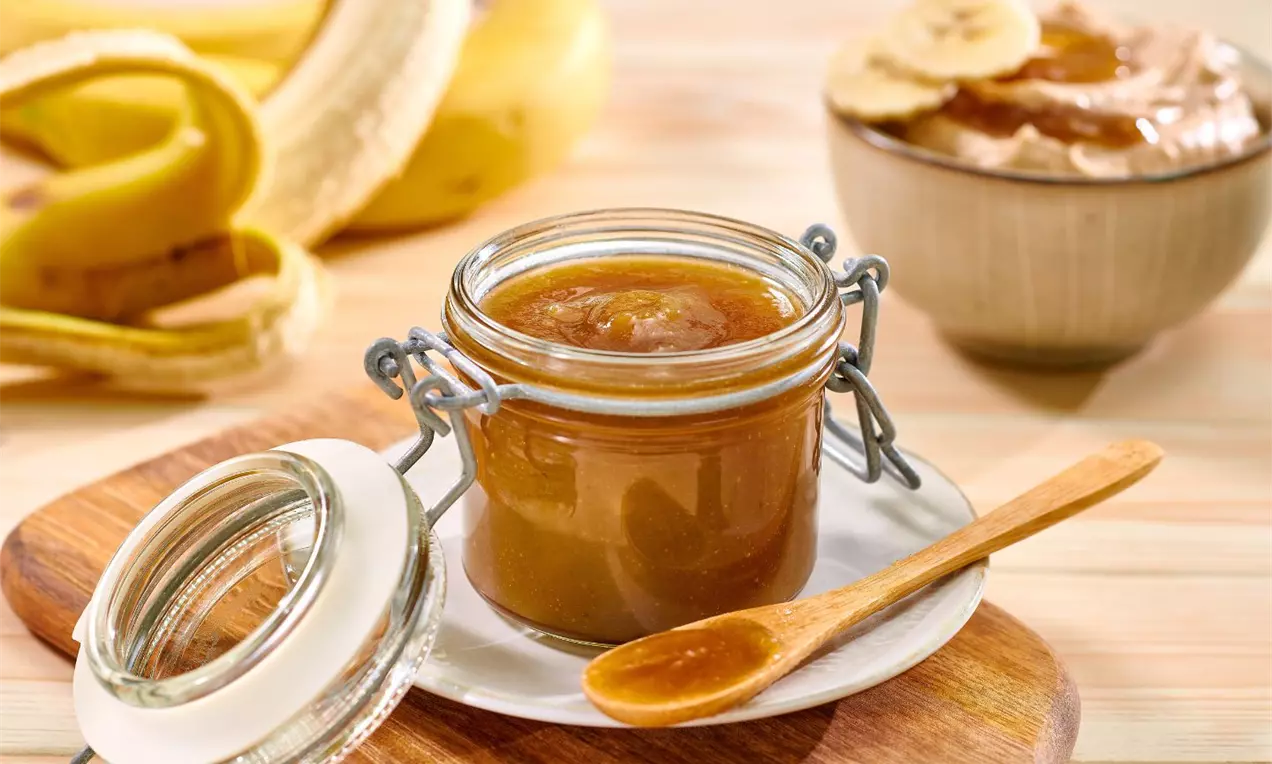 Cremă caramel aurie și lucioasă într-un borcan de sticlă transparent cu capac, așezată pe un tocător de lemn lângă o linguriță și felii de banană